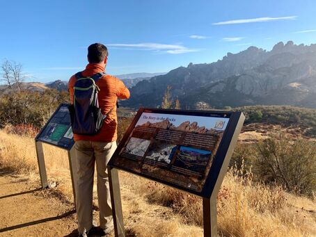 A man stands between two wayside educational signs looking toward the Pinnacles rock formations in the distance